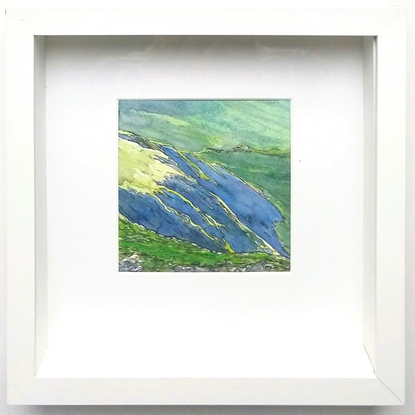 This is a framed view of "Crag Shadows" a 4.75 by 4.75 inch watercolor and ink painting on paper by Rebecca M. Fullerton. It depicts deep purple-blue shadows in a mountain ravine, with bright green slopes surrounding it. The painting is is a white mat inside a deep 10 by 10 inch shadowbox frame.