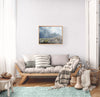 A framed landscape art painting hangs on a white wall above a light wood daybed with gray and white pillows and a striped throw blanket in a bright, modern living room.