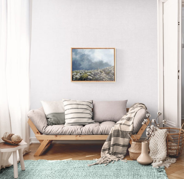 A framed landscape art painting hangs on a white wall above a light wood daybed with gray and white pillows and a striped throw blanket in a bright, modern living room.