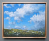 Framed painting of a blue sky with clouds and green landscape.