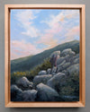 Painting of a person standing on rocky terrain with a scenic sky, framed in wood.