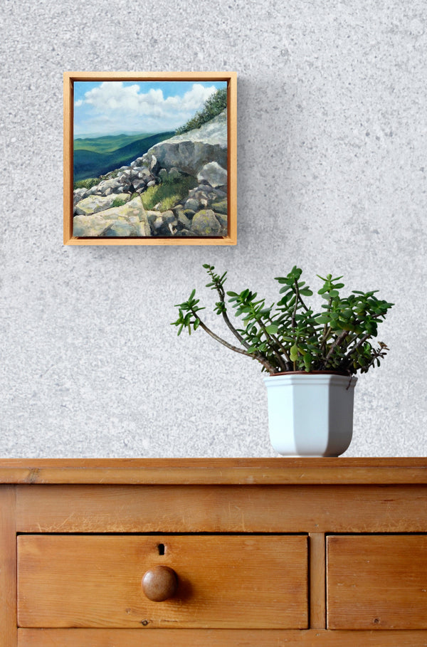 "A Sheltering Place" is a 10 by 10 inch oil on panel painting by Rebecca M. Fullerton, depicting a little grassy spot among the rocks on a mountain slope. Sunlight slants across the rocks and makes the spot look warm and inviting. Mountains and clouds fill the background. Seen here framed and hanging on a wall.
