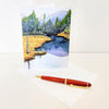 A large blank greeting card with a landscape painting depicting a stream and trees, accompanied by an envelope and a pen.