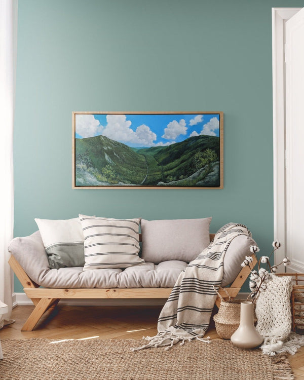 "Mount Willard, Summer" is a 20 by 40 inch framed oil on canvas painting by Rebecca M. Fullerton depicting the view over Crawford Notch from Mount Willard in the heart of New Hampshire's White Mountains. This artwork depicts mountains, valleys, forests, cliffs, trees, roads, railroad tracks, clouds, blue sky and summertime. This is the painting hung in a cozy living room.