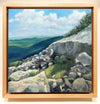 This is a framed view of "A Sheltering Place," a 10 by 10 inch oil on panel painting by Rebecca M. Fullerton, depicting a little grassy spot among the rocks on a mountain slope. Sunlight slants across the rocks and makes the spot look warm and inviting. Mountains and clouds fill the background.