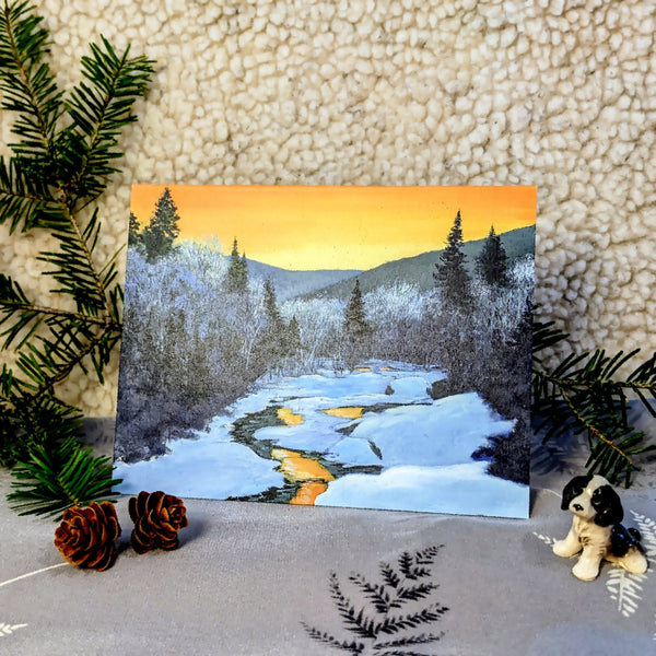 A small blank greeting card depicting a painted winter sunset scene with snow-covered trees and a river, accompanied by pine cones and a plush toy.